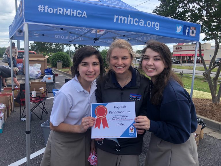 three girls smile for the camera. they stand in front of a tent with rmhca.org on one side