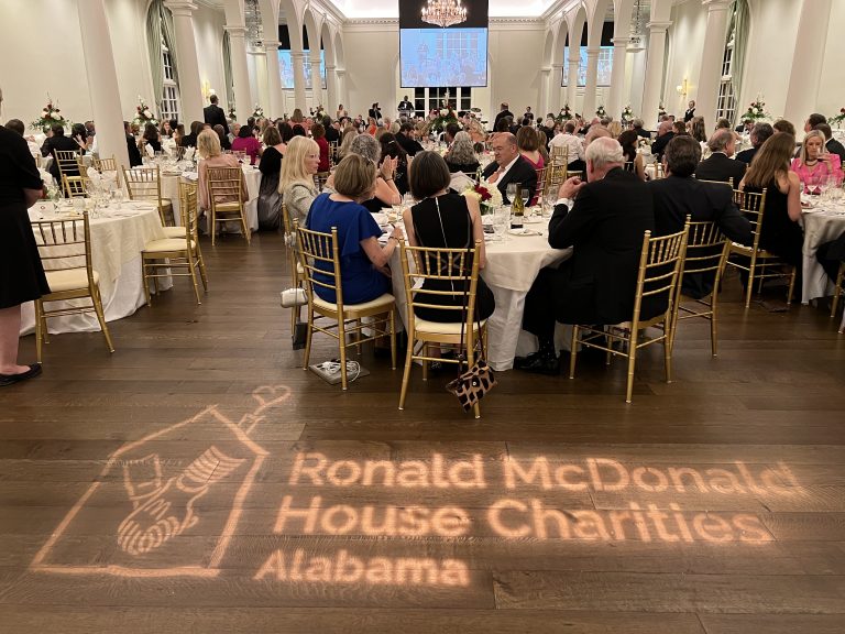 Gala guests sit at round tables. The floor beside them has a projected image of the RMHCA logo