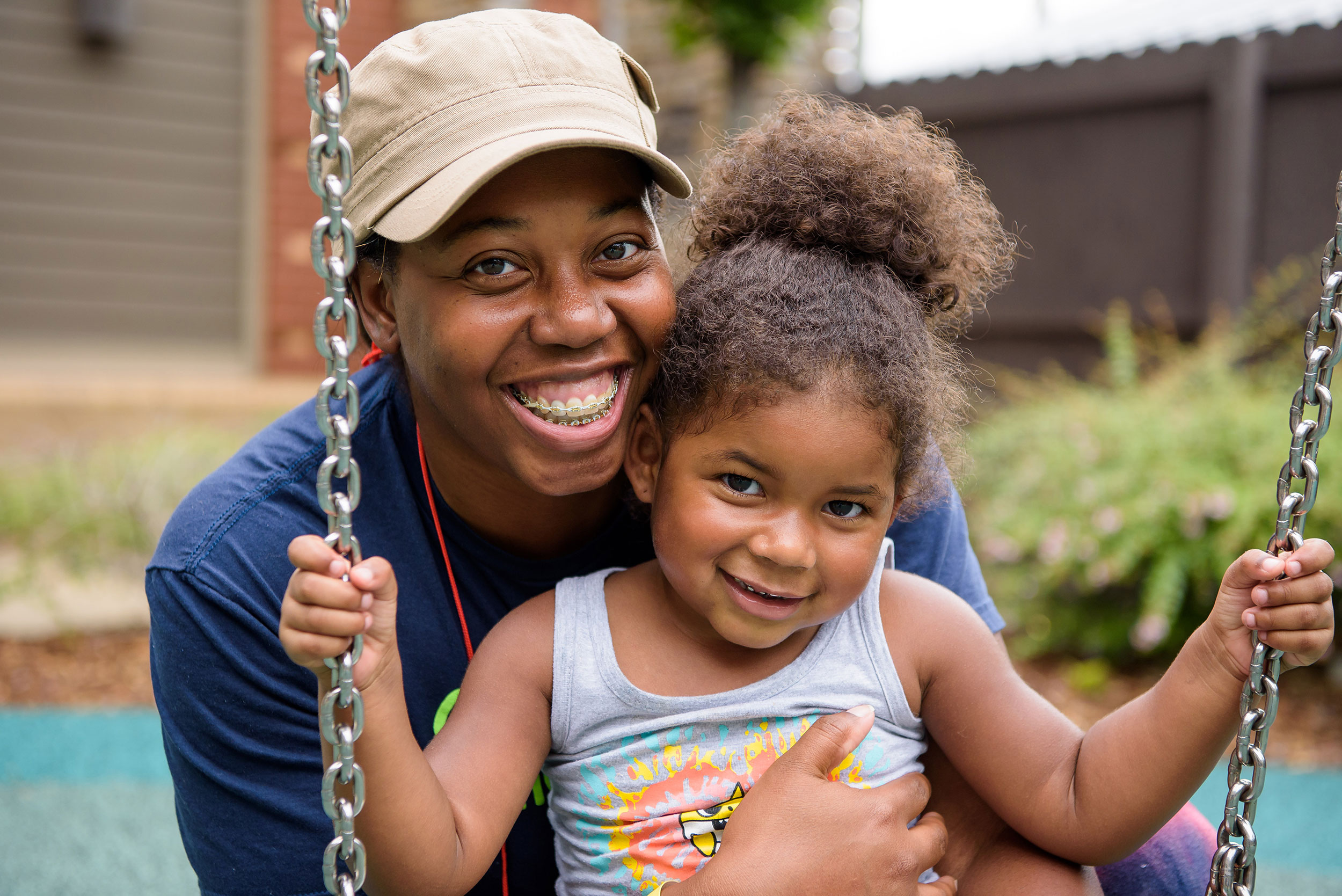 an older woman and a young girl pose for a photo. The young girl is sitting on a swing with the older woman's arms around her in a hug. They both are smiling.