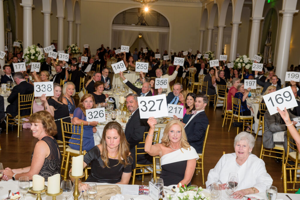 PEPOLE IN A BALL ROOM HOLDING UP AUCTION SIGNS.