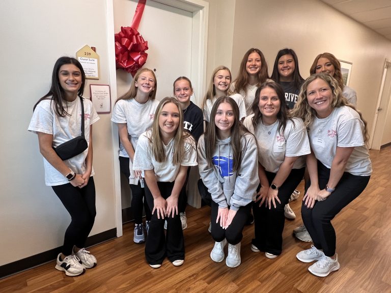 A group of students pose for a photo outside a room. The door is decorated with a red ribbon and bow